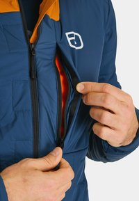 Blue puffer jacket with orange accents, featuring a front zipper pocket. The fabric appears smooth with a lightweight texture. Close-up on the zipper.