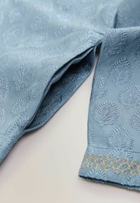 Blauwe textiel met een reliëfbladpatroon, met een gladde textuur en gouden geborduurde details langs de zoom.