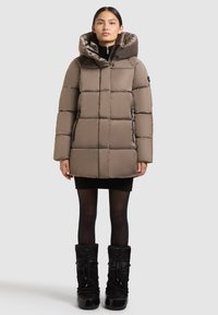 Woman wearing taupe padded hooded winter coat, black mini dress, sheer black tights, and black winter boots, standing against plain background.