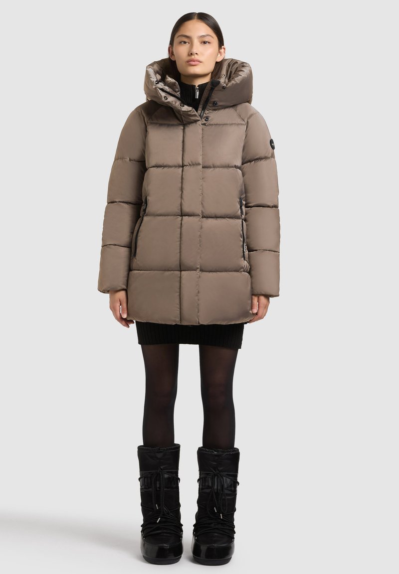 Woman wearing taupe padded hooded winter coat, black mini dress, sheer black tights, and black winter boots, standing against plain background.