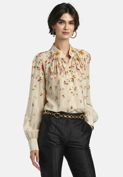 Woman wearing a cream floral blouse with long sleeves and black pants, accessorized with large hoop earrings and a gold chain belt.