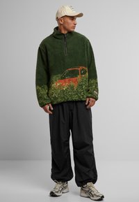 Grüner Fleece-Pullover mit halbem Reißverschluss, zeigt ein orangfarbenes Auto-Graphic zwischen dem Grün. Kombiniert mit lockeren schwarzen Hosen und Turnschuhen.