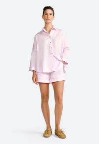 Chemise boutonnière rose clair à manches longues, accompagnée de shorts assortis. La tenue présente une coupe ample et un tissu lisse.