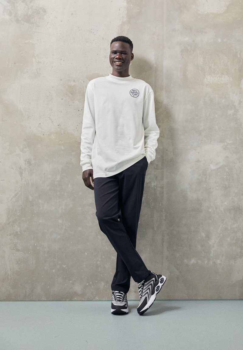 Camisa blanca de manga larga con un logo redondo, pantalones negros y zapatillas deportivas en blanco y negro con patrones ondulados, de pie frente a una pared gris.