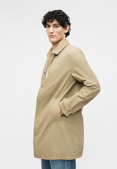 Νεαρός άνδρας με σγουρά μαλλιά, ντυμένος με μπεζ παλτό τύπου trench και μπλε τζιν, στέκεται με τα χέρια στις τσέπες μπροστά σε λευκό φόντο.