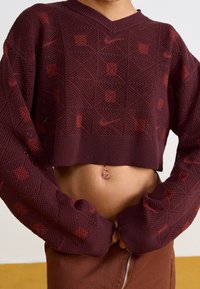 Burgundský zkrácený svetr s texturovaným pleteným vzorem, s výstřihem do V a dlouhými rukávy s čtvercovými akcenty v tmavším odstínu.