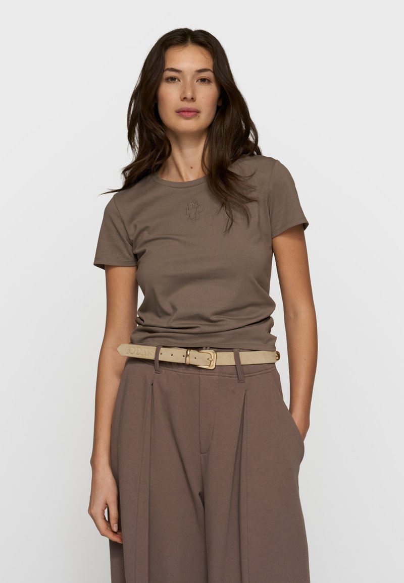 Femme aux longs cheveux bruns portant un t-shirt marron ajusté, un pantalon marron ample et une ceinture beige, debout devant un fond blanc uni.