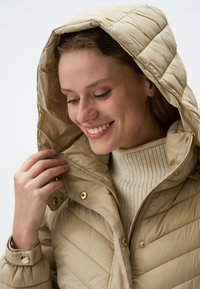 Beige gewatteerde jas met een capuchon, voorzien van diagonale quiltpatronen, gouden drukknoopsluitingen en een gestructureerde crèmekleurige trui eronder.