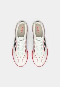 Λευκά ποδοσφαιρικά παπούτσια με μαύρες ρίγες και φωτεινές ροζ λεπτομέρειες, με λεία επιφάνεια και στρογγυλεμένο σχέδιο στη μύτη. Τα κορδόνια είναι λευκά.