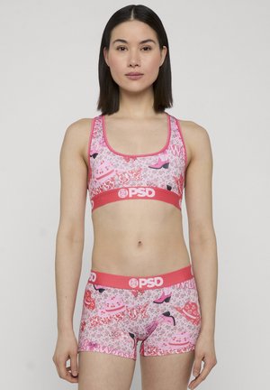 Mujer con un conjunto deportivo a juego en rosa con estampados de zapatos, sombreros, corazones y logotipos de "PSD" sobre un fondo liso.