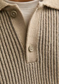 Beżowy i brązowy prążkowany sweter z dzianiny z dekoltem w serek i dwoma beżowymi guzikami, noszony na prostą beżową koszulę.