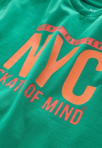 T-shirt en coton vert avec un grand graphique orange "NYC" au toucher lisse et un col ras du cou ; inclut "TWENTY FOUR SEVEN" au-dessus.