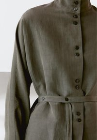 Diep olijfgroene linnen jurk met lange mouwen, knoopsluiting aan de voorkant, hoge kraag en bijpassende riem in de taille.