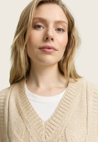 Un gilet en tricot beige avec un design en V, une texture côtelée et des motifs en relief, superposé sur une chemise blanche ajustée. Léger et décontracté.