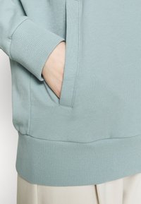 Sudadera de color azul claro con puños y cintura acanalados. Presenta un bolsillo lateral, textura suave y un diseño casual. Sin patrones visibles.