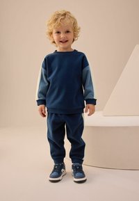 Granatowy sweatshirt z jasnoniebieskimi rękawami, zestawiony z pasującymi spodniami. Charakteryzuje się miękką teksturą i ściągaczami na rękawach. Noszony z niebieskimi sneakersami.