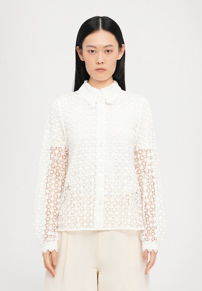 Blusa bianca in pizzo con motivo floreale testurizzato, chiusura frontale con bottoni e colletto. Presenta maniche lunghe e trasparenti e orlo ondulato.