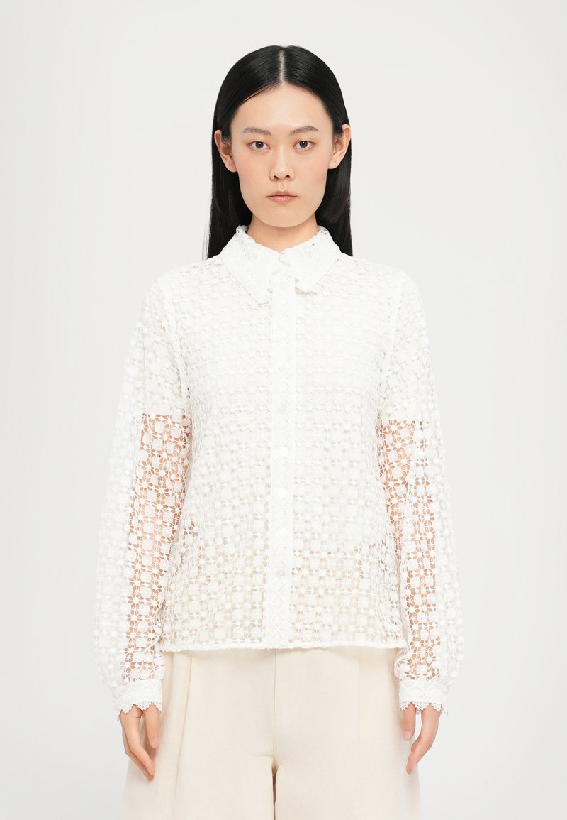 White lace blouse with a textured floral pattern, button-up front, and collar. Features long, sheer sleeves and a scalloped hem.