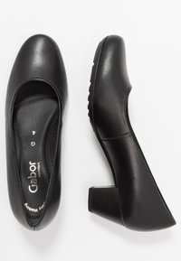 Svarta skinnklackar med rund tå, texturerad klack och perforerad innersula. Varumärkesnamnet tryckt på insidan av sulan.