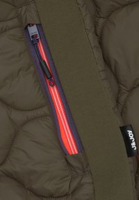 Donkergroen gequilt stof heeft een rode rits met een zwarte trekband en een groene gebreide rand. Een label met witte tekst bevindt zich aan de zijkant.