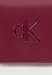 Hlboká burgundská kožená plocha s vyvýšeným lesklým logom "cK" a viditeľným šitím pozdĺž dolného okraja.