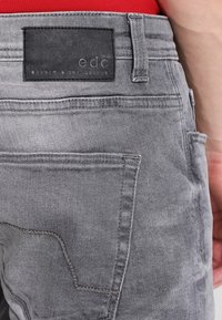 Los jeans de mezclilla gris presentan una etiqueta de cuero con la marca "edc" en la parte trasera, mostrando un diseño de cinco bolsillos y una textura lavada.