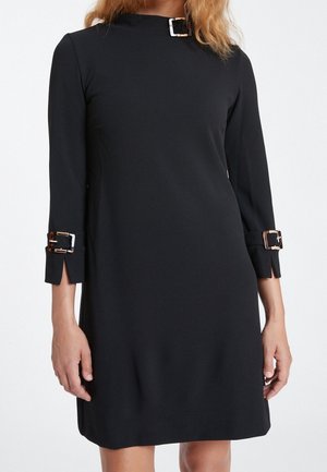Woman wearing a black knee-length dress with long sleeves, featuring tortoiseshell buckle details at the collar and cuffs.
