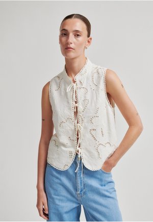 Kvinne med tilbakeglattet hår iført ermeløs hvit brodert topp med knyting foran og lyseblå jeans, stående med hånden i lommen.