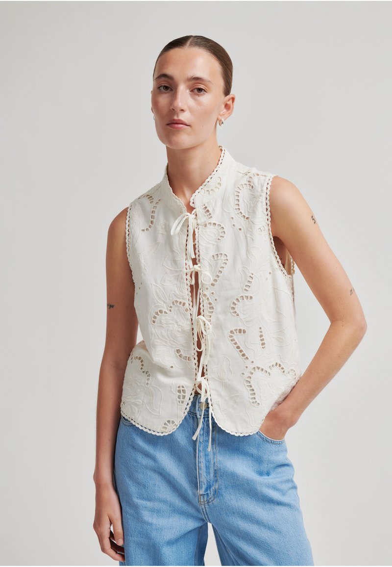 Femme aux cheveux plaqués en arrière portant un top blanc brodé sans manches avec des liens à l'avant et un jean bleu clair, debout, main dans la poche.