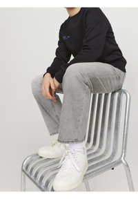 Osoba v šedých džínách, bílých teniskách a černé mikině sedí se spojenýma rukama na stříbrné kovové židli s horizontálními pruty.