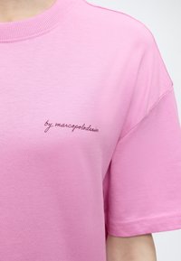 Κοντινό πλάνο ενός ροζ κοντομάνικου πουκάμισου με την επιγραφή "by marcopolodenim" κεντημένη με μικρά καλλιγραφικά γράμματα στην αριστερή πλευρά του στήθους.