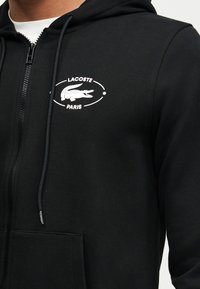 Hoodie noir zippé avec logo Lacoste Paris blanc comprenant un crocodile sur le côté gauche de la poitrine, porté sur une chemise blanche.