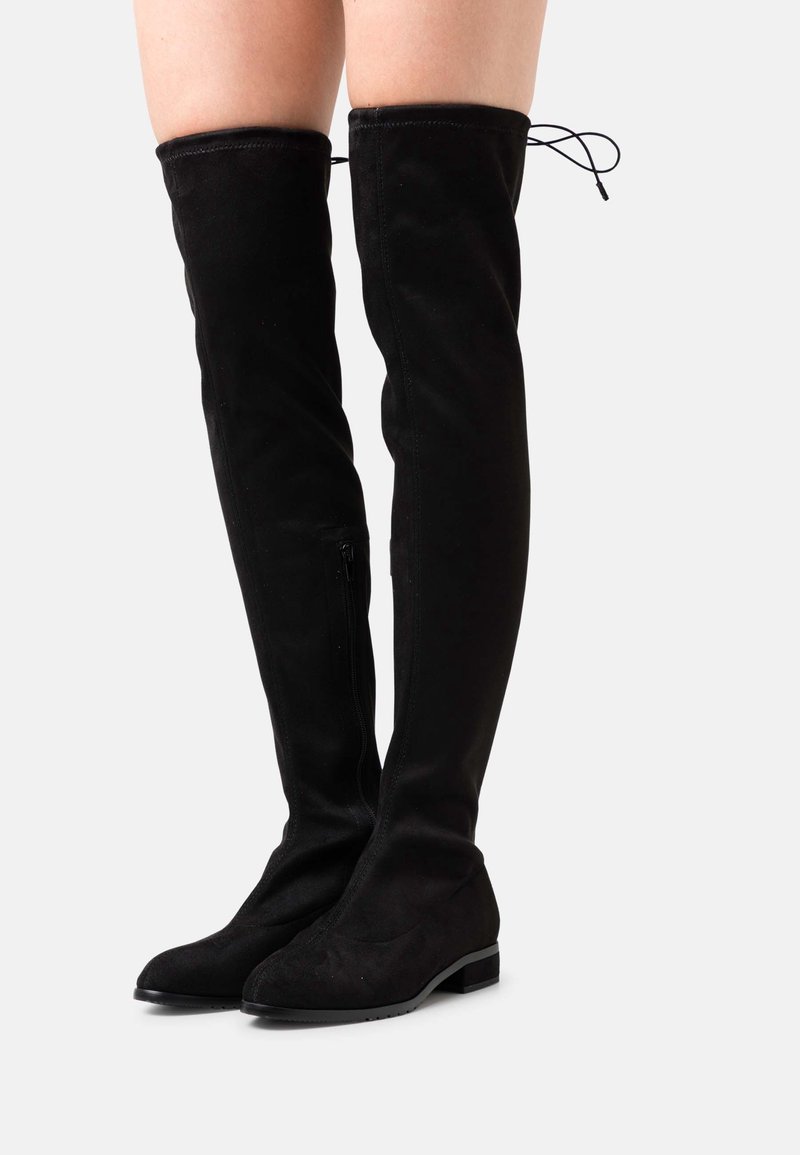 Bottes plates en daim noir montant au-dessus du genou, avec un petit talon bloc et un détail de lien à l'arrière, portées sur des jambes nues sur un fond uni.