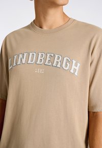 Vaaleanbeige puuvillapaita, jossa on pyöreä pääntie. Paidassa on valkoisella ja sinisellä brodeerattu "LINDBERGH" vuonna "1987". Lyhyet hihat.