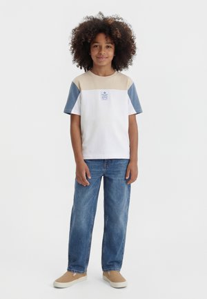 Persona joven con cabello rizado que lleva una camisa de manga corta bloqueada en colores beige, blanco y azul, vaqueros azules y zapatos beige sin cordones.