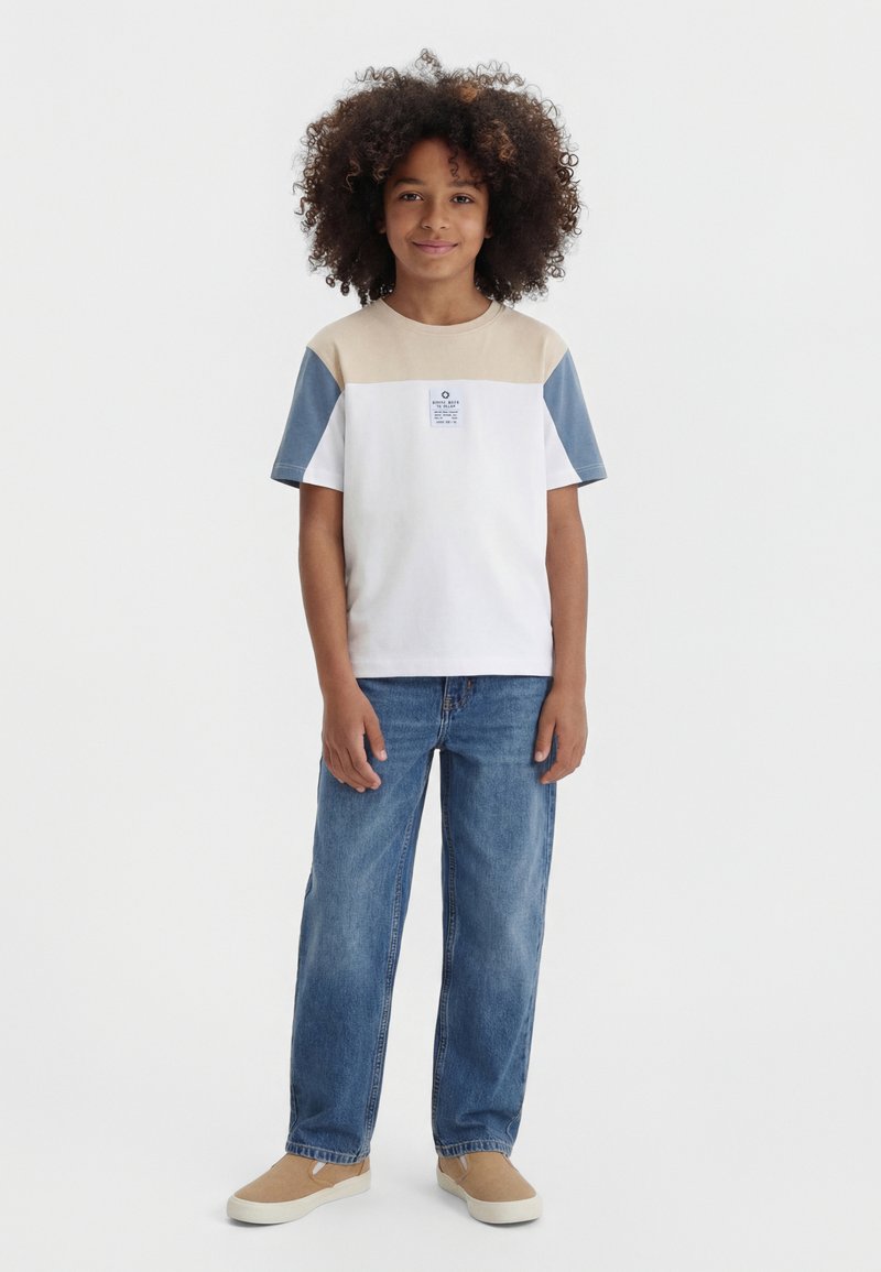 Mladá osoba s kučeravými vlasmi, ktorá nosí tričko s krátkym rukávom v béžovej, bielej a modrej farbe, modré džínsy a béžové slip-on topánky.