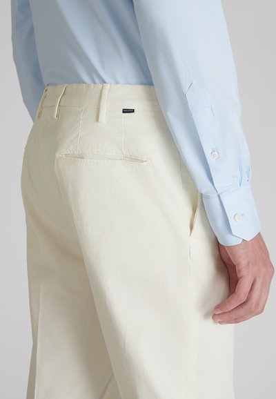 Pantaloni beige chiaro con una texture liscia, dotati di una tasca posteriore e un branding discreto; abbinati a una camicia a maniche lunghe azzurro chiaro.