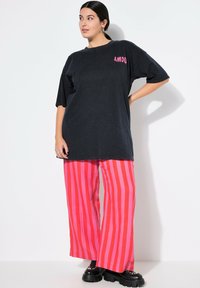 Zwarte oversized t-shirt met "AMOU" in het roze op de linkerkant van de borst, gecombineerd met wijde roze en rode gestreepte broek en chunky zwarte schoenen.
