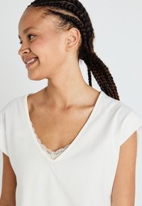 Top bianco a V con maniche corte, bordo in pizzo attorno al collo e vestibilità rilassata. Tessuto morbido con una superficie liscia.