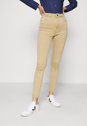 Personne portant un pantalon beige ajusté, des baskets blanches avec des détails noirs, et une chemise à manches longues bleu marine, les mains sur les hanches.