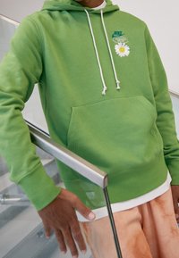 Zelená mikina s kapucí, která má klokánkovou kapsu, bílé stahovací šňůrky a grafikou sedmikrásky s logem. Nositelná s kalhotami s jemným tanovým batikovaným vzorem.