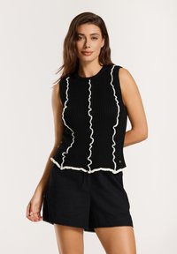 Must have mustra- ja pealöige ilma varrukateta musta kõrge kvaliteediga kudumi top, mille servades on valged volangid ja vertikaalsed jooned; sobitatud mustade lühikeste pükstega.