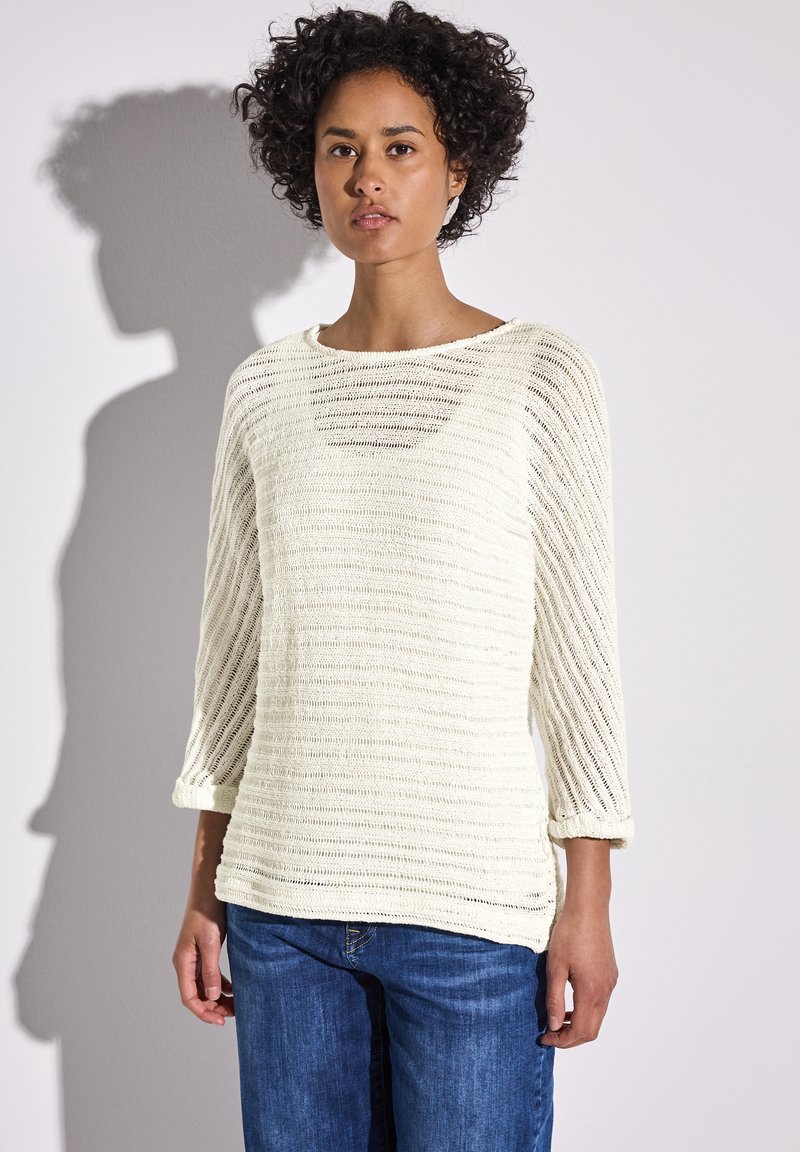 Hellecremiger gestrickter Pullover mit horizontalen Streifen, Bootsausschnitt und dreiviertellangen Ärmeln. Kombiniert mit blauen Jeans.