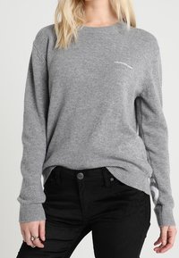 Grå stickad tröja med långa ärmar, rund halsringning och ribbade mudd. Litet vitt logotyp på bröstet. I kombination med svarta jeans.