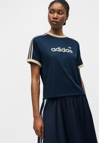 Žena v tmavě modrém sportovním oděvu Adidas s béžovým lemováním a logem, stojící před jednoduchým světlým pozadím, hledící stranou.