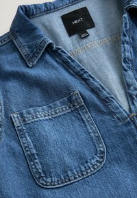 Denim-paita, jossa on V-pääntie, lyhyt kaulus ja yksi rintatasku. Sinisessä kankaassa on hienovarainen tekstuuri ja kontrastiväriset ompeluyksityiskohdat.