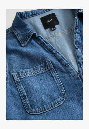 Chemise en denim avec un col en V, un col court et une poche poitrine unique. Tissu bleu affichant une texture subtile et des détails de couture contrastants.