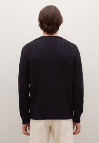 Persona con capelli corti castani, che indossa un maglione scuro a trecce e pantaloni chiari, in piedi di fronte a una parete leggera e semplice.