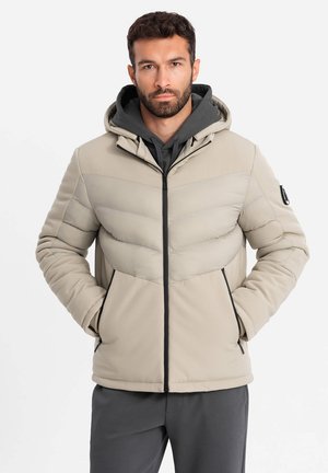 Lichtbeige pufferjack met een ritssluiting aan de voorkant, een capuchon en contrasterende donkere accenten. Heeft een chevronontwerp en zijzakken.