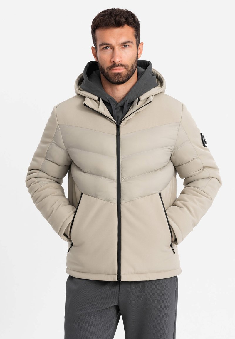 Giacca puffer beige chiaro con zip frontale, cappuccio e accentuazioni scure a contrasto. Presenta un design a zigzag e tasche laterali.